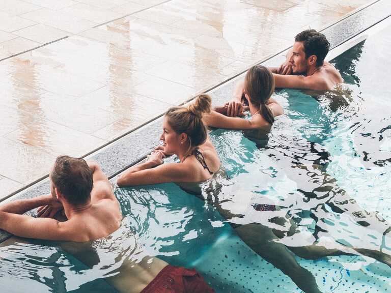 Gäste im Innenpool des Hotel Eibl-Brunner. Abgestützt am Beckenrand entspannen Sie im warmen Wasser des Pools.