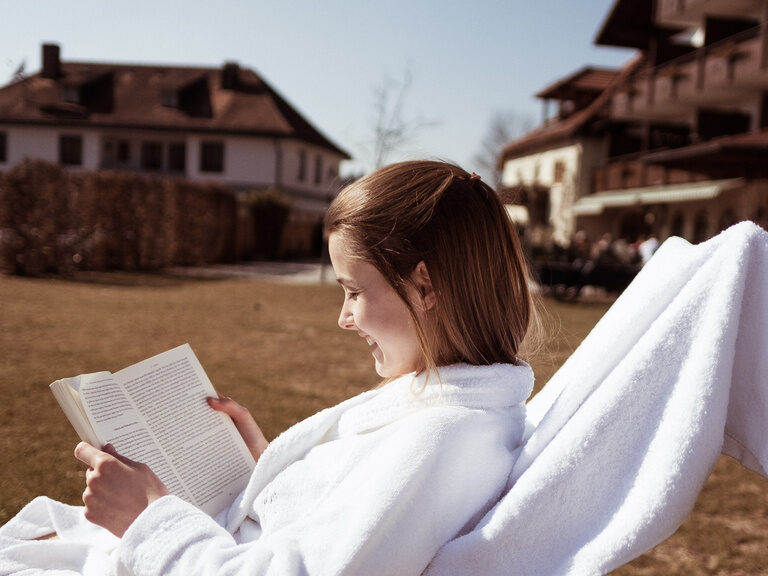 Frau genießt die Sonnenstrahlen, auf der Sonnenliege, im Hotelgarten des Wellnesshotel Eibl-Brunner, beim Lesen eines Buches.
