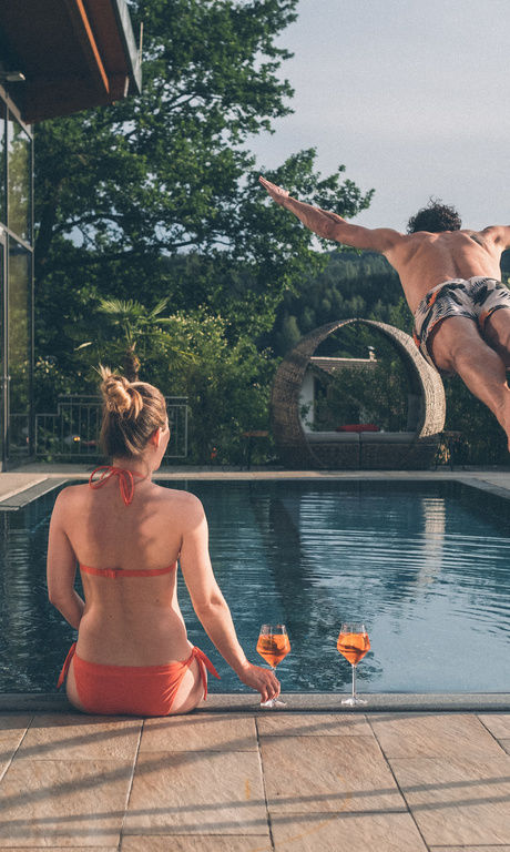 Ein Paar am Pool genießt die Sonne. Die Frau sitzt am Poolrand mit einem Glas Aperol und schaut dem Mann bei einem Sprung in das Wasser zu.