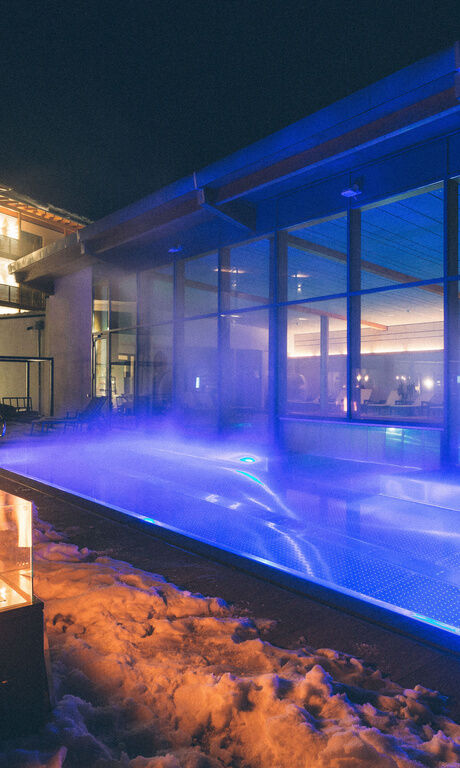 Verschneites Hotel und Garten mit dampfenden Außenpool und Ambientebeleuchtung, sowie Feuerstellen für romantisches Licht.