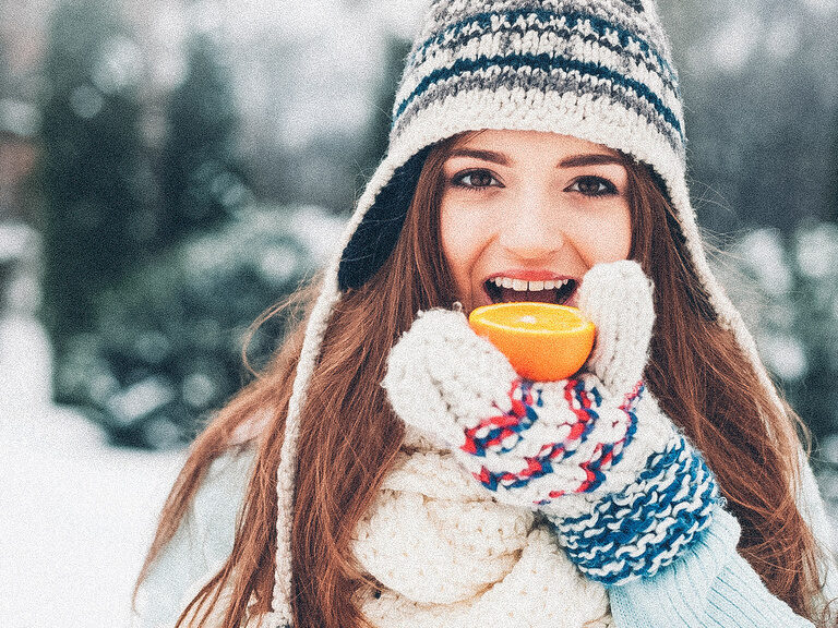 Mit Mütze und Handschuhen bekleidet beisst eine Frau in eine Orange.