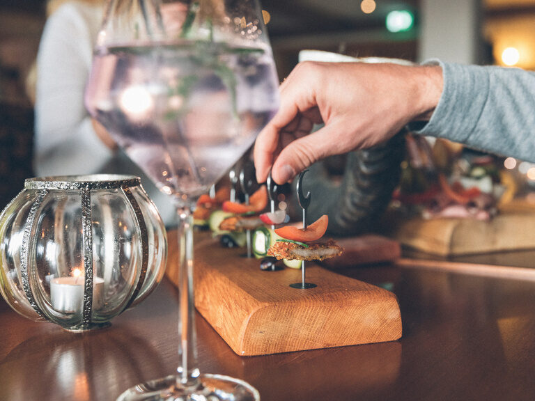 Fingerfood auf Spiesen werden zwei Gästen/innen gereicht. Gemeinsam mit zwei Getränken aus der Bar des Hotels.