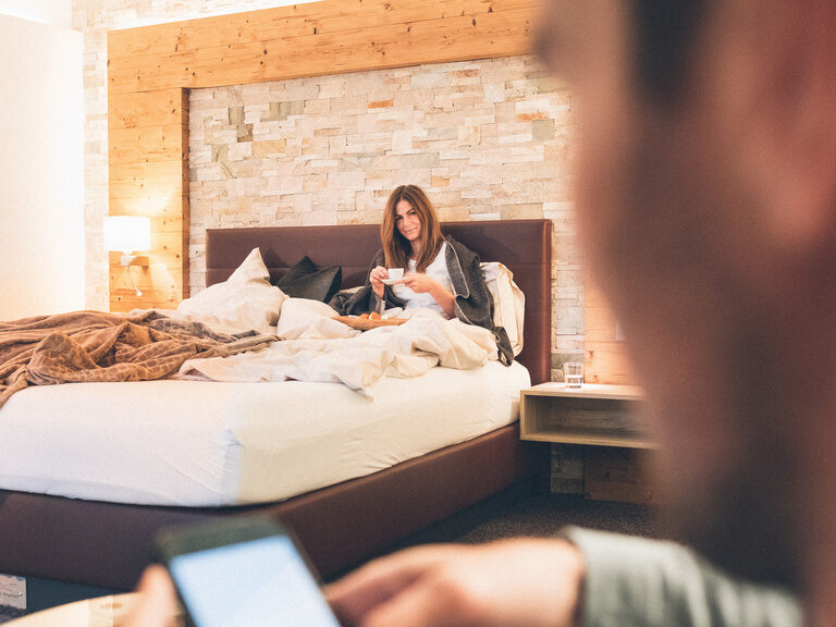 Gäste im Doppelzimmer des Hotel Eibl-Brunner. Die Frau genießt Ihren Kaffee im Bett und der Mann schaut auf sein Handy.