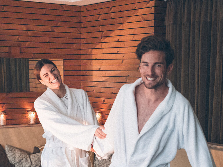 Ein Gästepaar geht, mit Bademänteln bekleidet, aus dem Ruhebereich mit den gemütlichen Entspannungsliegen in die Saunawelt des Hotel Eibl-Brunner.