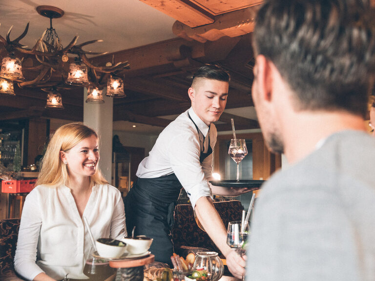 Lächelnde Gästin wird von einem Kellner am Tisch bedient und bekommt ein Getränk serviert.