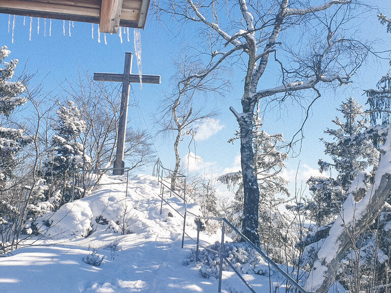 Verschneite Anhöhe mit Gipfelkreut, mit einem schmalen Weg zum Kreuz.