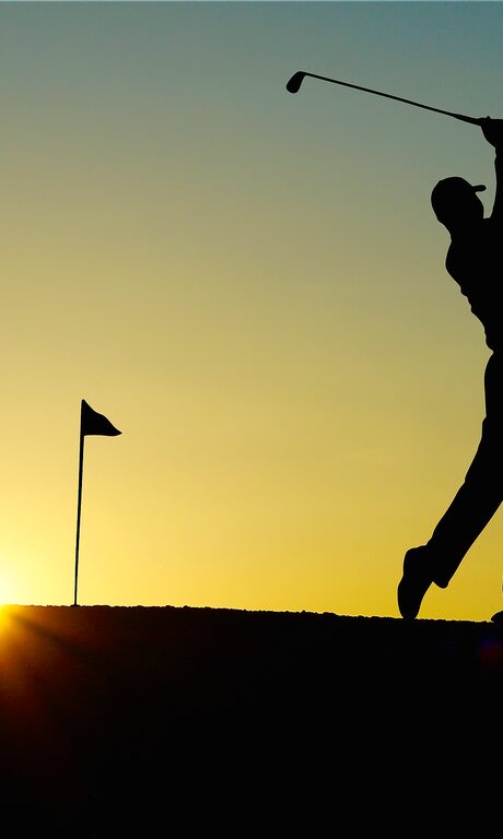 Golfer beim Schlag eines Golfballs, die Fahne im Rücken bei Sonnenuntergang.