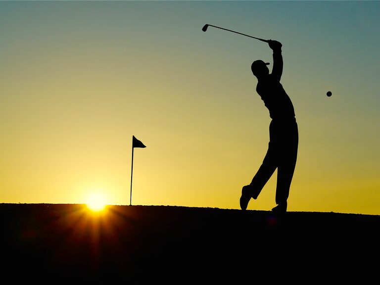 Golfer beim Schlag eines Golfballs, die Fahne im Rücken bei Sonnenuntergang.