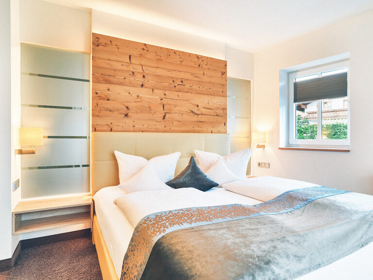 Doppelbett im Hotel Eibl-Brunner, in einem hellen Zimmer mit Fenster und Holzverkleidung am Kopfteil des Bettes.