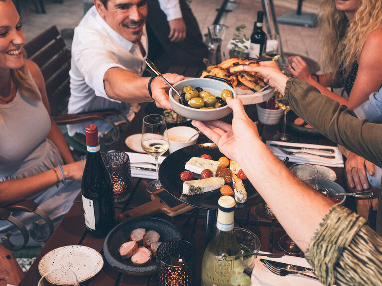 Reichgedeckter Tisch mit mediterranen Speisen, einer Flasche Wein und Personen die sich gegenseitig das Essen reichen und teilen.
