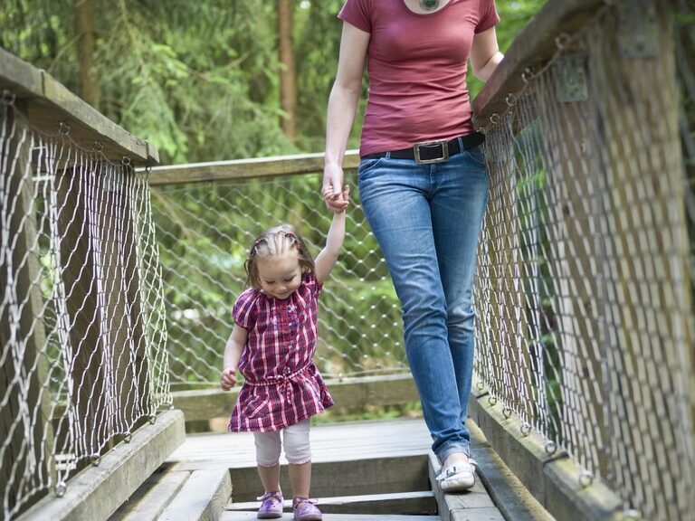 Eine Mutter balanciert mit ihrem Kind an der Hand, umgeben von grünen Bäumen, über den Balancierpfad im Baumwipfelpfad Nationalpark Bayerischer Wald.
