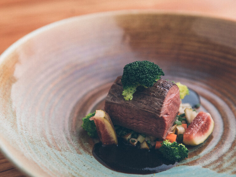 Nahaufnahme eines kurzgebratenen, rosa Rindfleisches, auf in Brunoise geschnittenen Wurzelgemüse, mit einer Jus und Feigen.