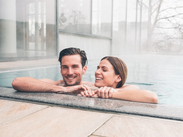 Ein Paar genießt die Temperaturen des warmen, dampfenden Außenpools des Hotel Eibl-Brunner, am Beckenrand abgestützt.