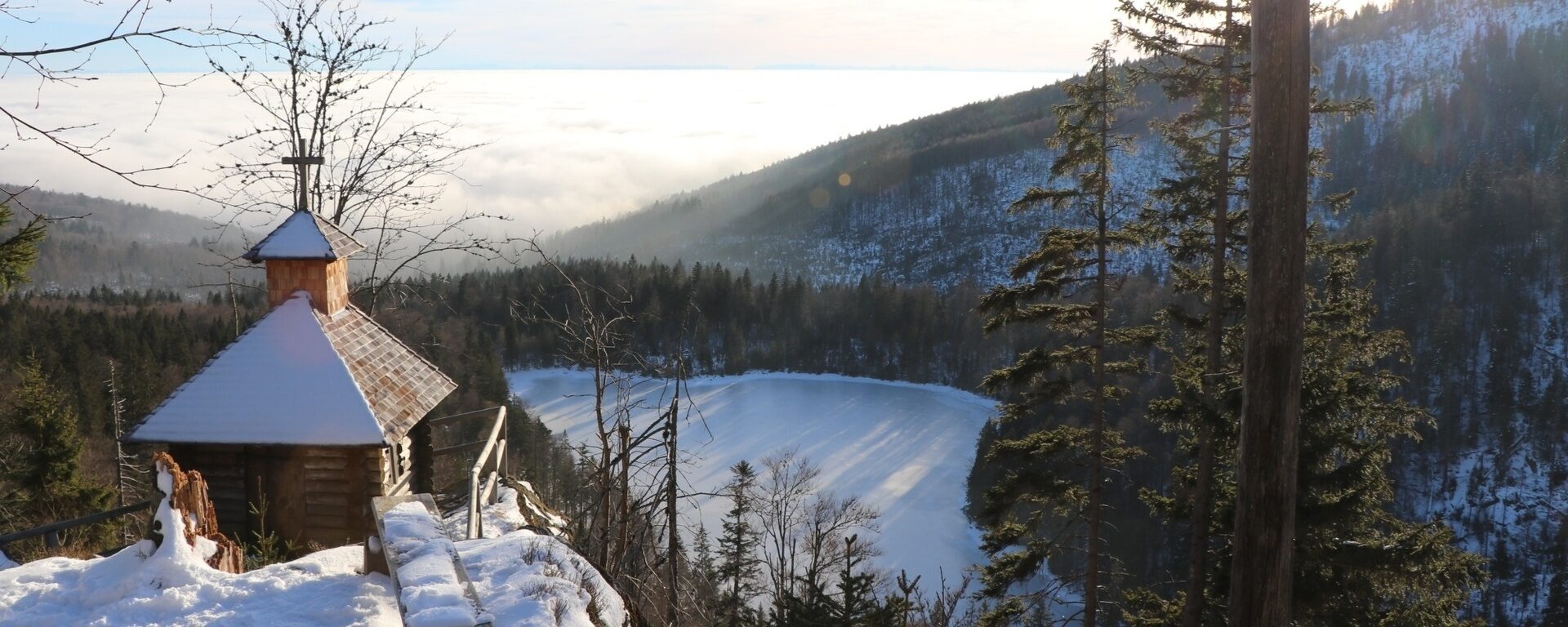 Mit Blick auf den gefrorenen Rachelsee steht man auf einer verschneiten Anhöhe in der Nähe der Kapelle. Im Hintergund liegt dichter Nebel.