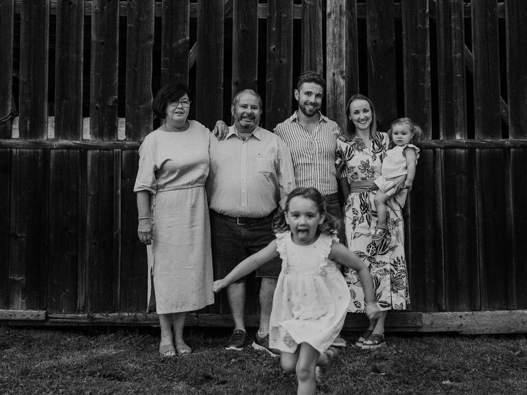 Fröhlich lächelnde Familie Brunner mit Kindern vor einer Holzwand in schwarz-weiß.
