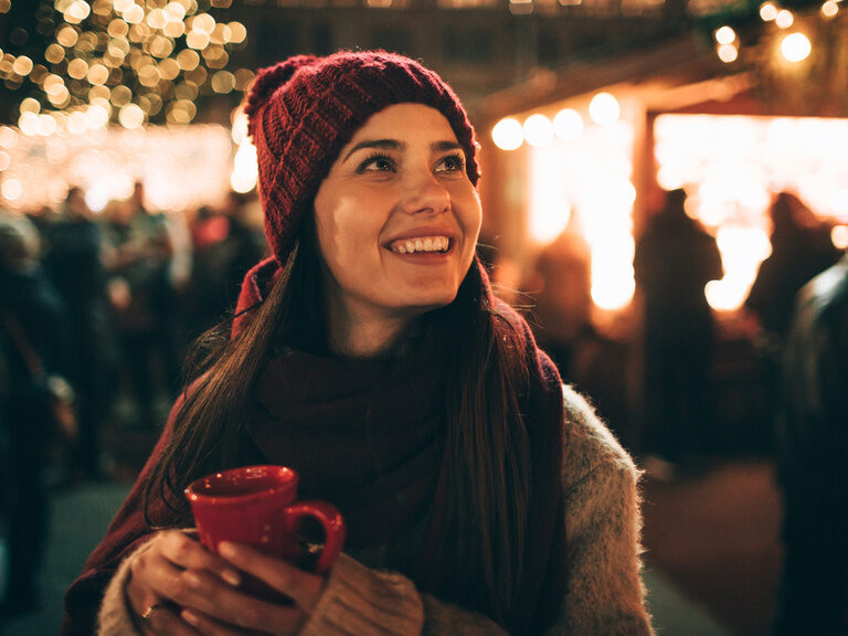 Frau erfreut sich an Lichtern und der Stimmung des Weihnachtsmarkts. Warm angezogen hält sie ein heißes Getränk in der Hand.
