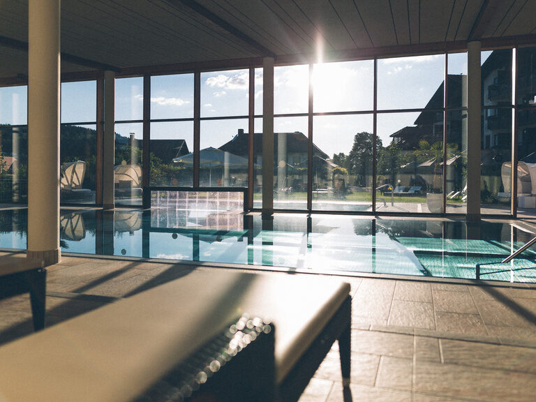 Gemütliche Liegen mit Blick durch die Fensterfront in den Hotelgarten und die Sonnenstrahlen erhellen die Wellnesslandschaft.
