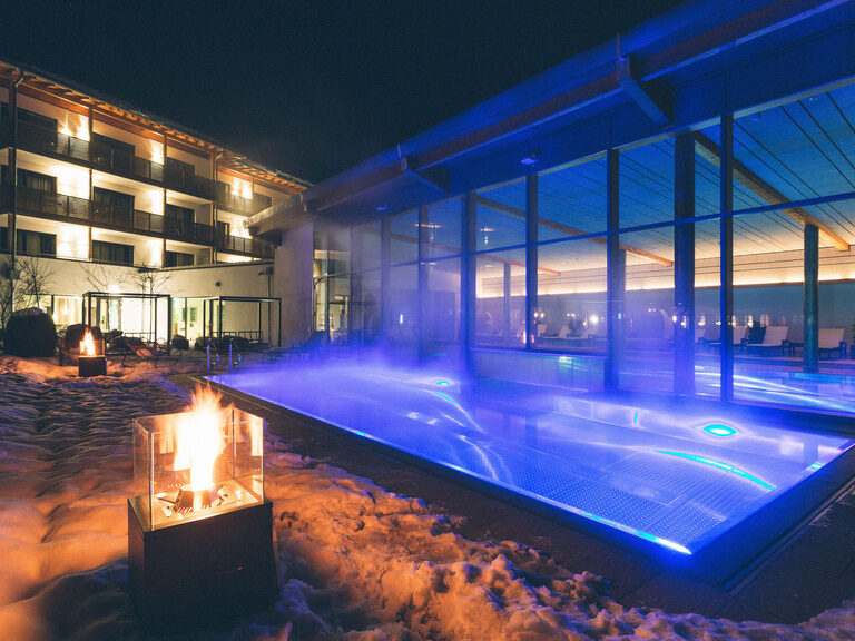 Verschneites Hotel und Garten mit dampfenden Außenpool und Ambientebeleuchtung, sowie Feuerstellen für romantisches Licht.
