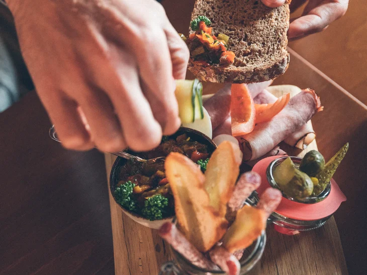 Hände belegen ein Brot mit Aufstrich aus einer Holzplatte mit Wurst und Gemüse