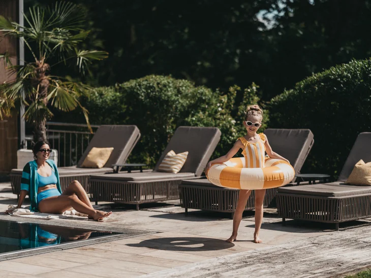 Kind mit Schwimmring und Frau in Badebekleidung am Pool mit Liegestühlen