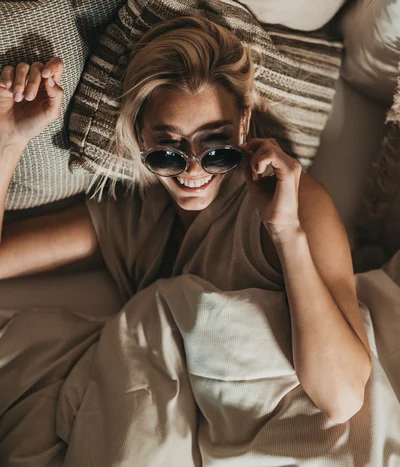 Lächende Frau liegt im Bett und richtet ihre Sonnenbrille mit Kissen um sie