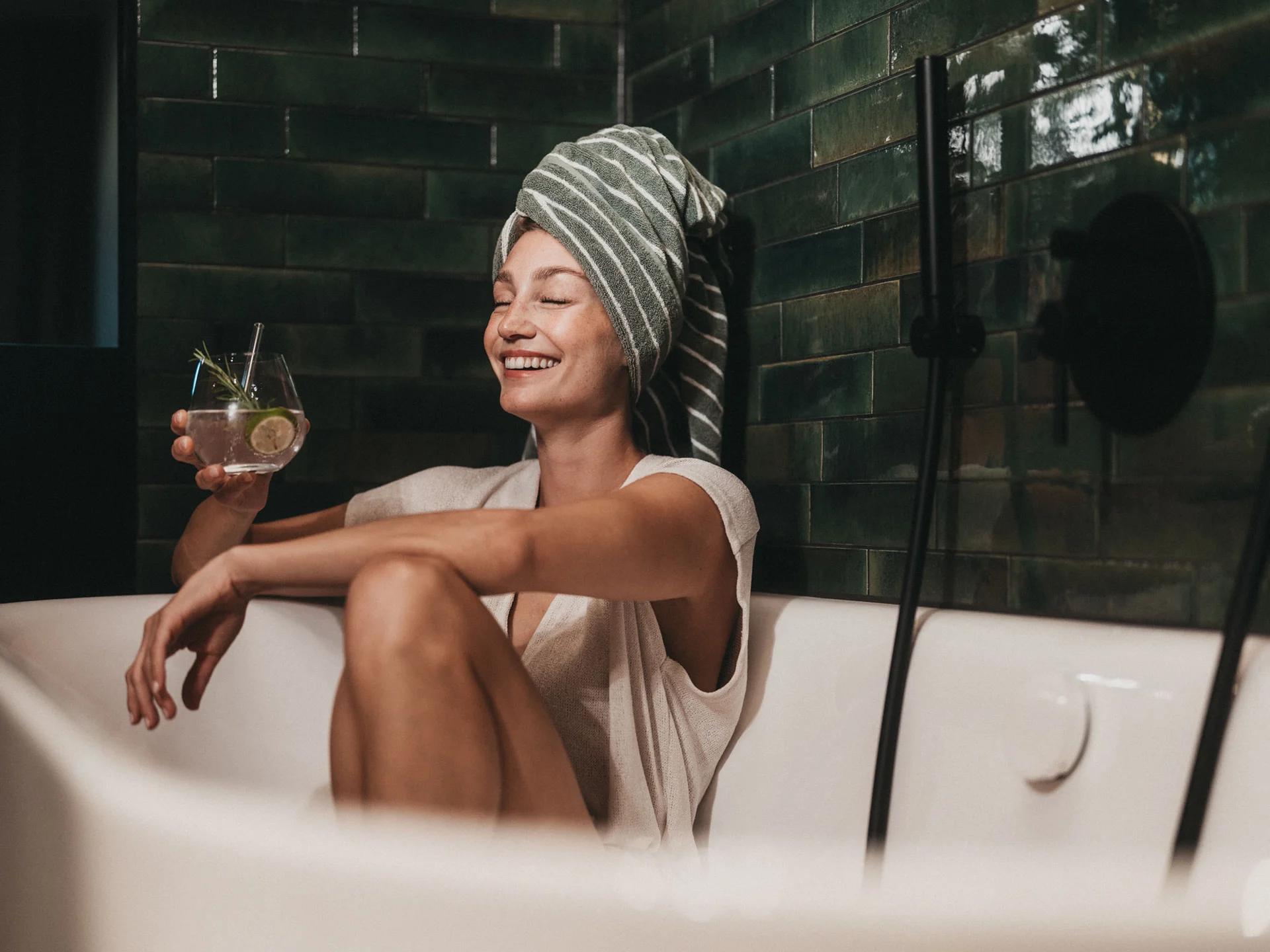 Lächende Frau mit Handtuch auf dem Kopf entspannt in der Badewanne mit Getränk