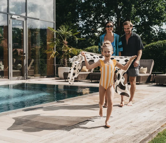 Kind im gestreiften Badeanzug läuft mit Tuch am Pool, Eltern folgen lächelnd