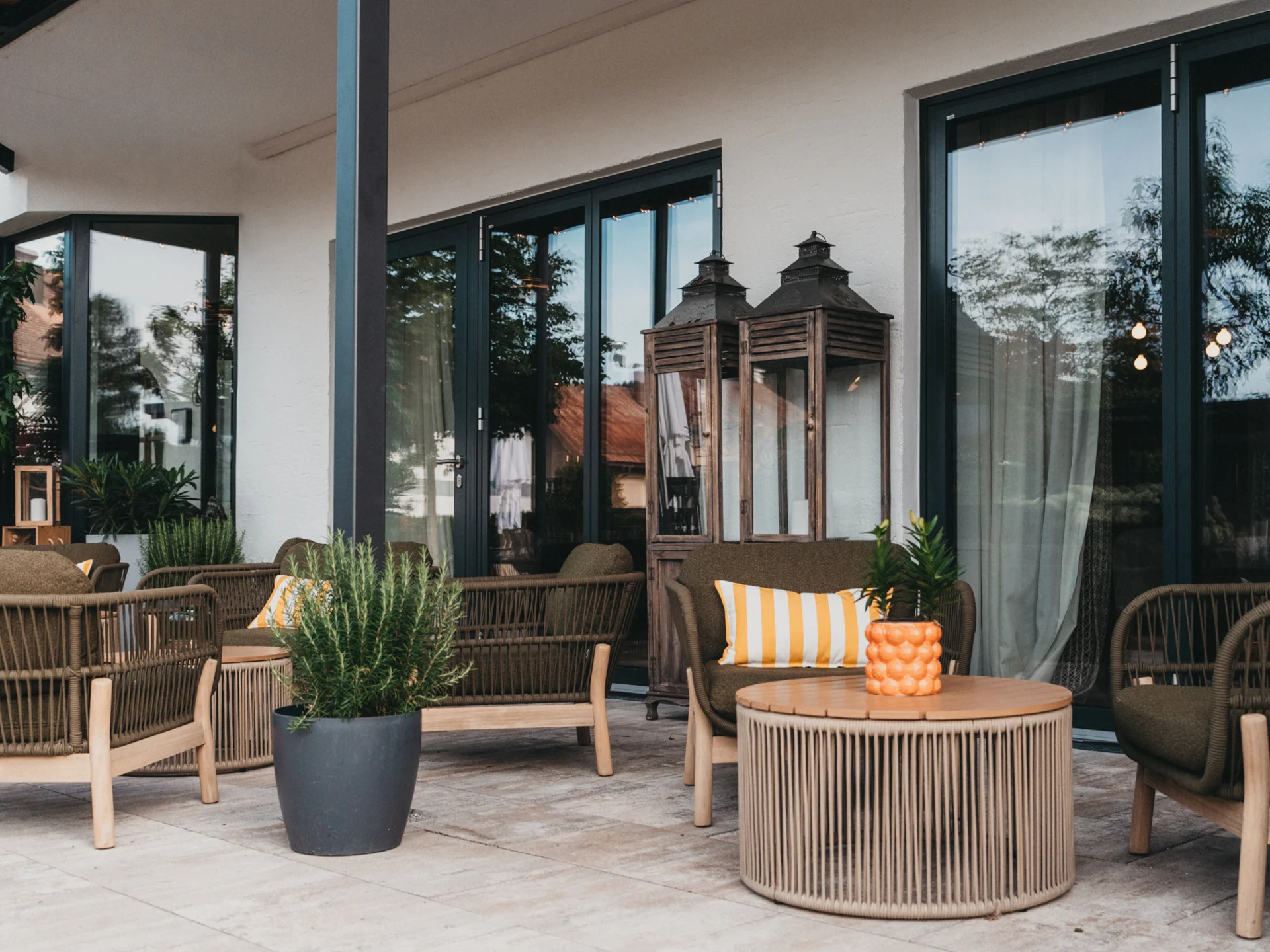 Gemütliche Terrasse mit Sitzmöbeln, Pflanzen und großen Laternen vor Glasfenstern