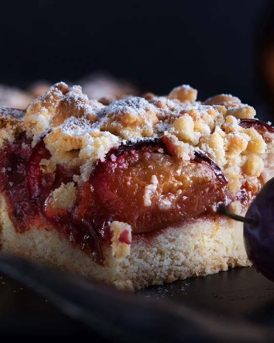 Ein Stück Pflaumenkuchen mit Streuseln und frischen Pflaumen auf schwarzem Teller