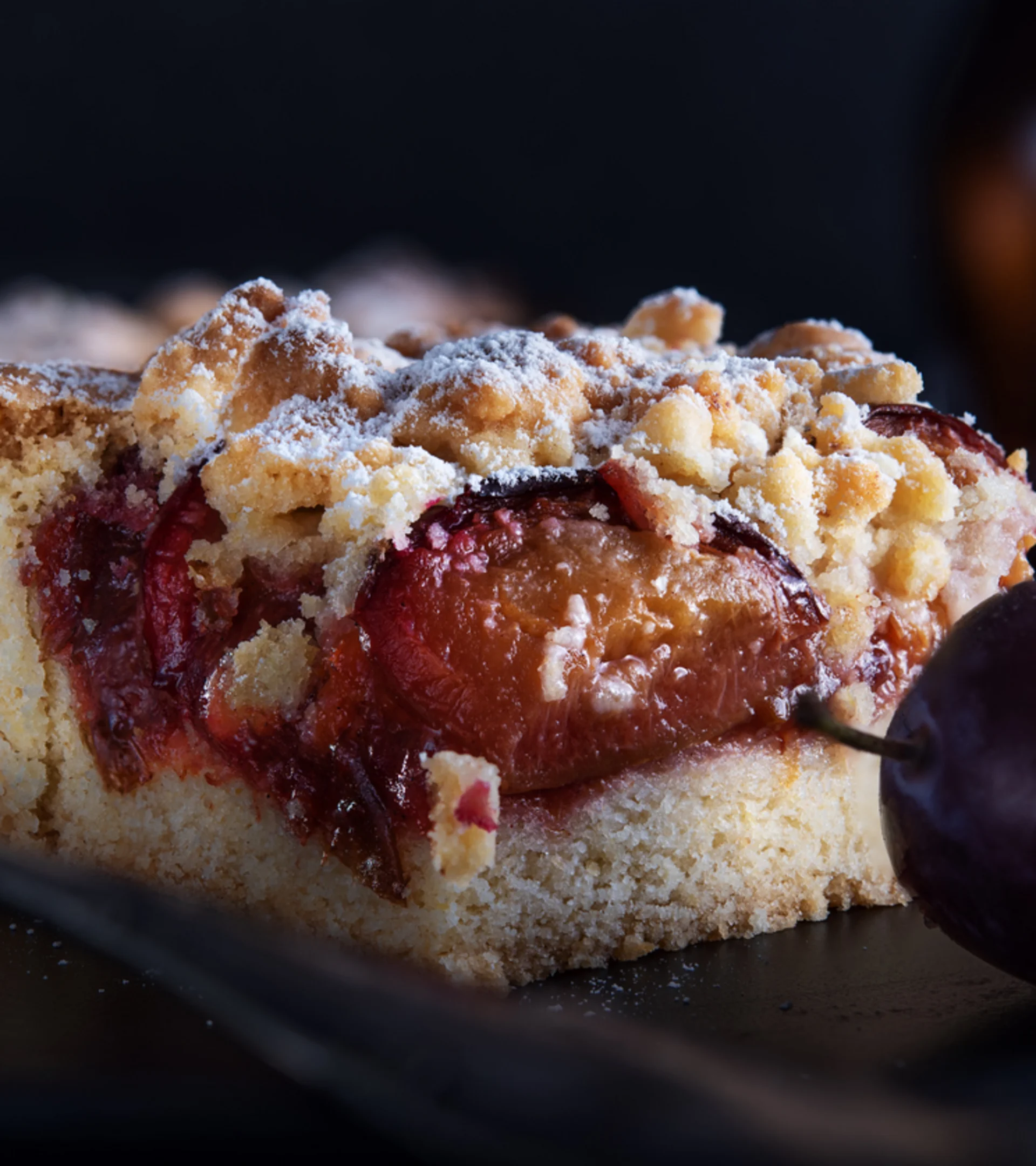 Ein Stück Pflaumenkuchen mit Streuseln und frischen Pflaumen auf schwarzem Teller